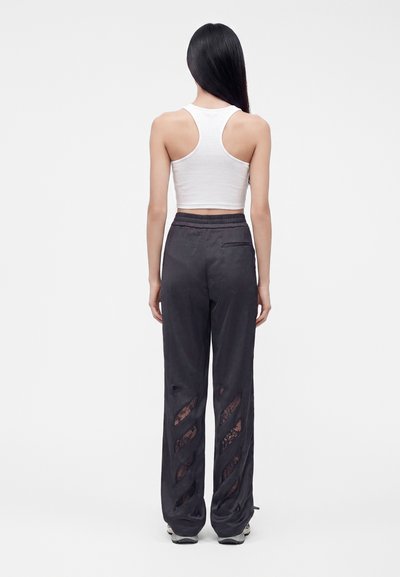Femme aux longs cheveux noirs portant un crop top blanc sans manches et un pantalon noir ample avec des découpes en mesh, vue de dos.
