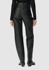Achteraanzicht van een persoon in hoge zwarte leren broek, zwarte geribbelde lange mouw top en zwarte enkellaarsjes met hakken.