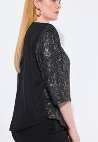 Sort bluse med trekvartærmer, der har et pailletdesign i nuancer af blå og guld på den ene side, og en glat tekstur på den anden.