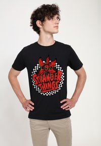 Czarny T-shirt z czerwonym nadrukiem przedstawiającym tekst "Stranger Things" otoczony wzorem w kratę. Miękka bawełniana tkanina, dopasowany krój.