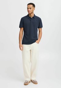 Tumši zila polo krekls ar īsām piedurknēm un apkakli, apvienots ar krēmkrāsas brīviem biksēm un pelēkiem sporta apaviem ar baltajiem šņorēm.