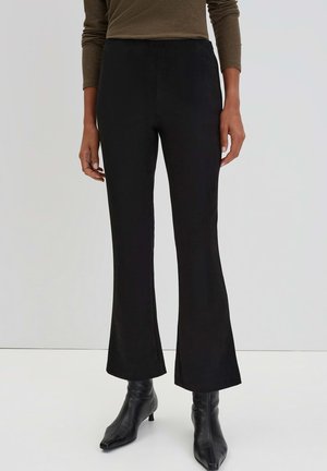 Schwarze, ausgestellte Hosen aus glattem Stoff, mit hohem Bund und leichtem Glanz. Kombiniert mit enganliegenden schwarzen Stiefeln bis zur Knöchel.