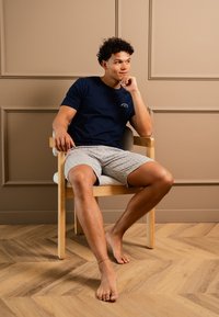 Marinblå crew-neck t-shirt med en liten logotyp, ihopparad med ljusgrå shorts med prickat mönster. Sitter på en trästol, barfota.