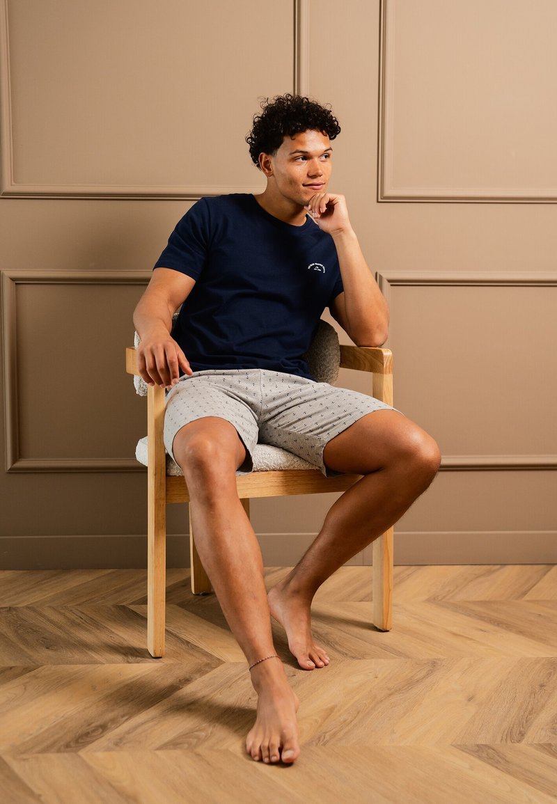Marinblå crew-neck t-shirt med en liten logotyp, ihopparad med ljusgrå shorts med prickat mönster. Sitter på en trästol, barfota.