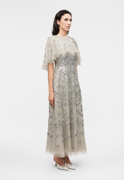 Γυναίκα με μακρύ, μπεζ φόρεμα με ασημένιες παγιέτες και κεντήματα με λουλούδια, διάφανα, ριγωτά μανίκια και λευκά μυτερά τακούνια, στέκεται πλάγια.