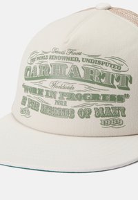 Kreminės spalvos kepurė su plokščiu kraštu, su tekstūruotu audiniu ir tinkliniu nugarėle. Žalia spausdinta tekstas, įskaitant "Carhartt" ir "Work In Progress."