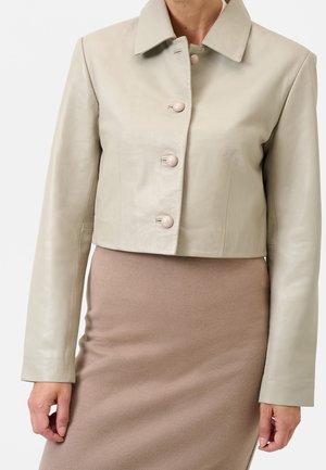 Giacca corta in pelle beige con grandi bottoni rotondi indossata sopra una gonna in maglia taupe aderente, mostrata dal collo ai fianchi.
