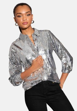 Silberne Paillettenbluse mit Kragen, Knopfschluss und Dreiviertelärmeln. Strukturierten Oberfläche reflektiert Licht. Kombiniert mit schwarzen Jeans.