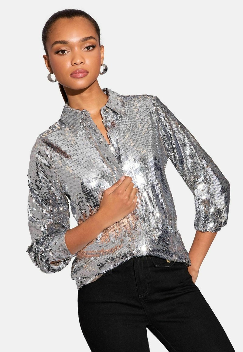Silver sequin blouse with a collared neck, button closure, and three-quarter sleeves. Textured surface reflects light. Paired with black jeans.