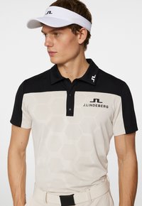 Beige och svart polo skjorta med ett hexagonmönster, med krage, tre knappar och en logotyp på bröstet och axeln.