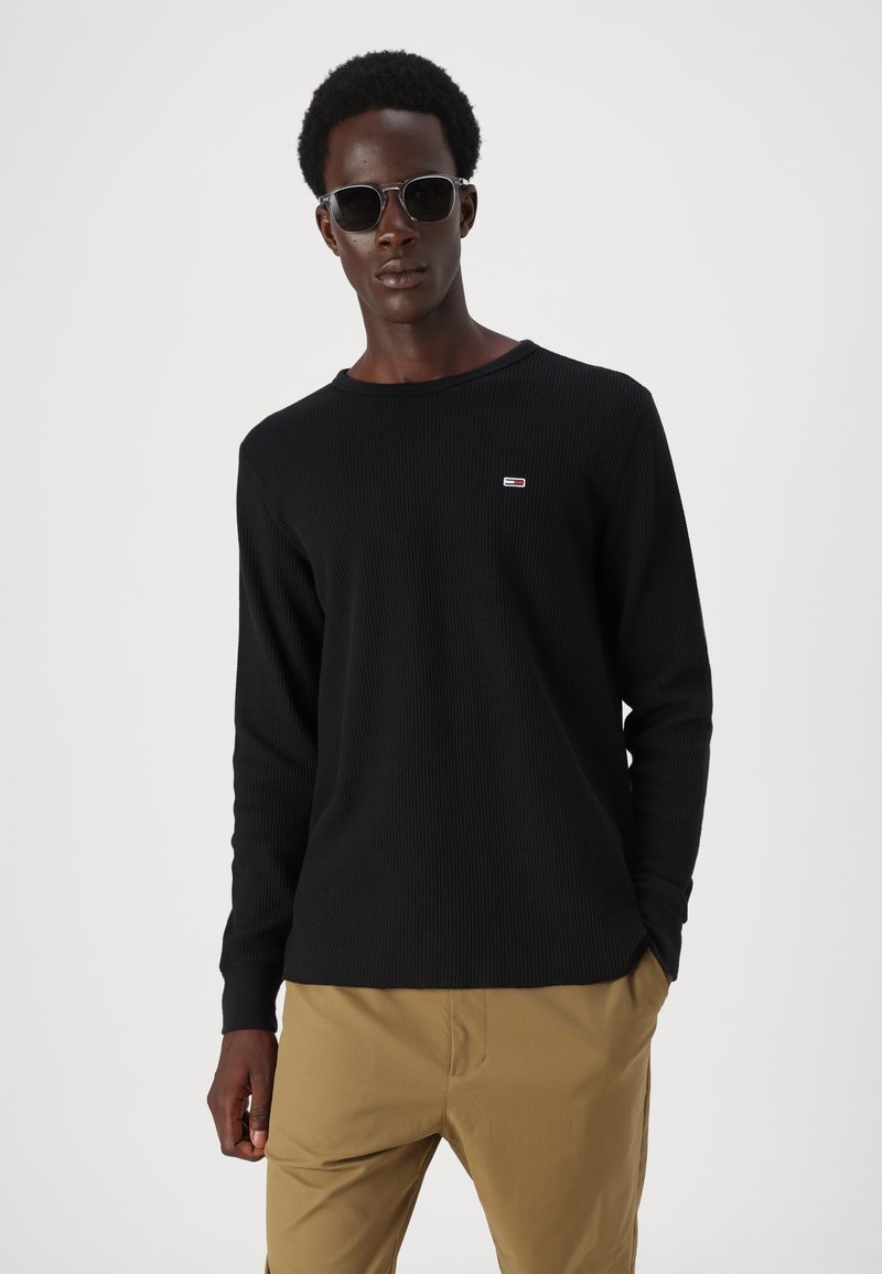 Czarny długi rękaw top z fakturowanym wzorem i okrągłym dekoltem, z małym detalem logo na klatce piersiowej, w połączeniu z khaki spodniami.