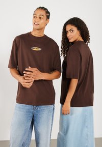T-shirts marron à manches courtes avec un logo blanc et orange sur la poitrine, portés avec un pantalon en jean bleu clair et une jupe. Coupe simple et décontractée.