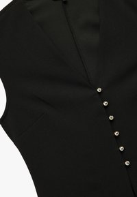 Sort vest med dyb V-udskæring, der har en enkelt brystlomme og seks metalliske knapper ned langs fronten. Glat stoftekstur.