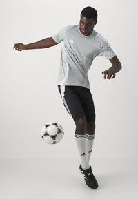 Szürke sportpóló fekete sorttal, fehér díszítéssel, fekete focicipővel és csíkos szürke zoknival párosítva; a focilabda mozgásban.