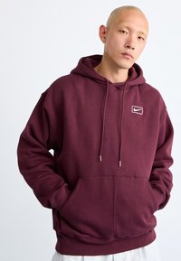 Maroon hooded sweatshirt made of soft fleece fabric. Features a front pocket and a small white Nike logo on the chest. Drawstring adjustable hood.