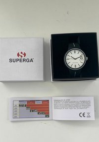 Reloj negro con una esfera texturizada blanca, acentos plateados y una correa de silicona, presentado en una caja de terciopelo negro, junto a un folleto de información del producto.