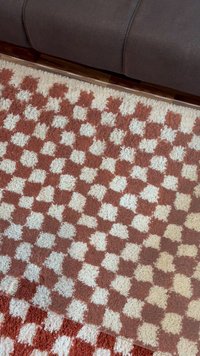 Tapete texturizado com um padrão xadrez nas cores ferrugem e creme. Fibras macias e felpudas criam uma superfície aconchegante. Borda próxima a um sofá escuro.