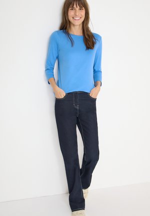 Blue long-sleeve top paired with dark denim jeans. The jeans feature contrasting stitching and a relaxed fit. Model stands with hands in pockets.