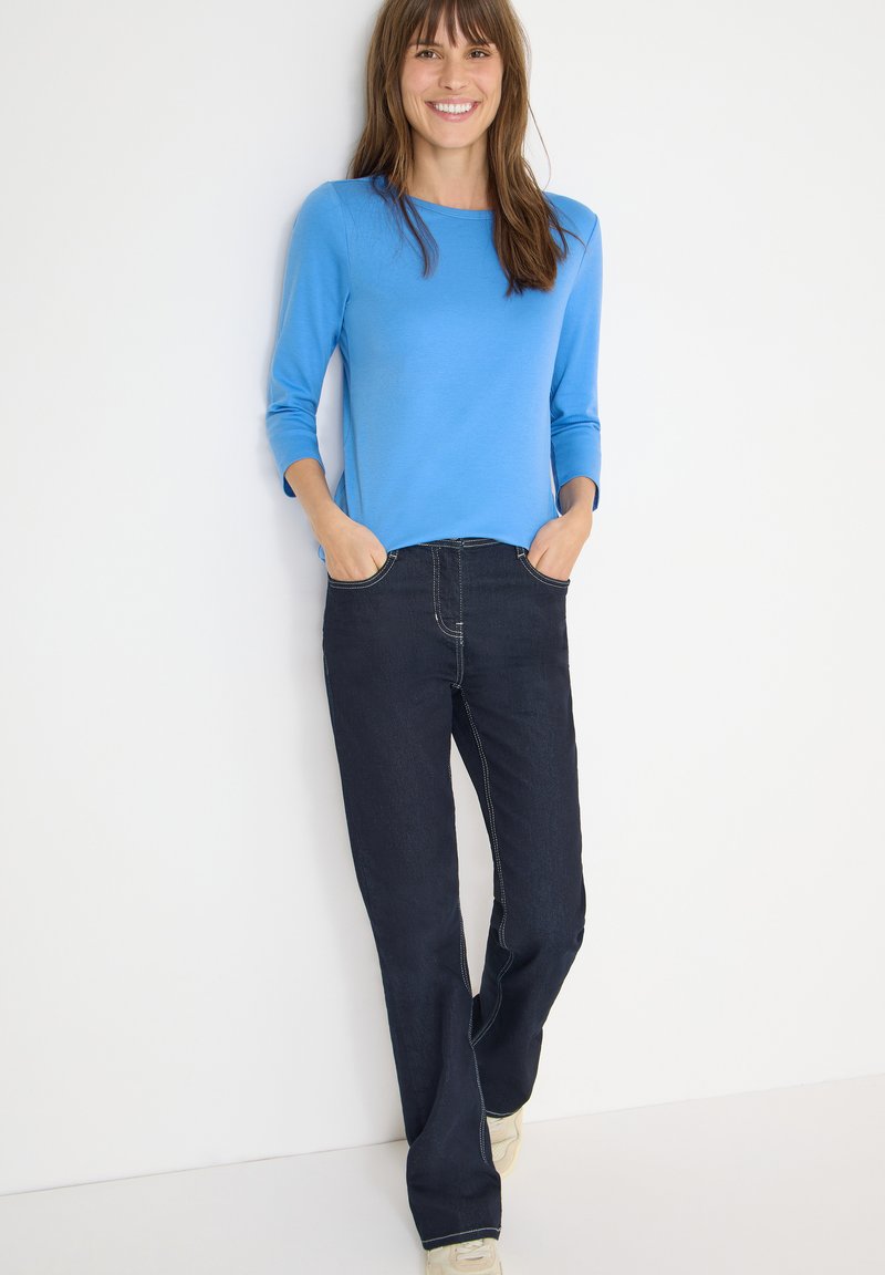 Blue long-sleeve top paired with dark denim jeans. The jeans feature contrasting stitching and a relaxed fit. Model stands with hands in pockets.