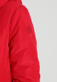 Veste rembourrée rouge avec une texture lisse, une capuche et un logo circulaire avec un "H" sur le haut de la manche.