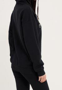 Felpa nera con vestibilità comoda, dotata di cappuccio con coulisse, polsini e orlo a coste. Dettaglio di design nella parte frontale, texture del tessuto morbida.