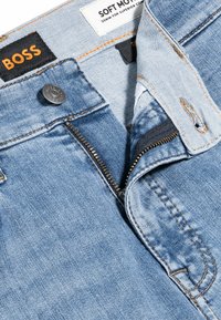Ljusblå denimjeans med dragkedja, orange sömmar och en svart etikett med "BOSS" och "SOFT MOTION".