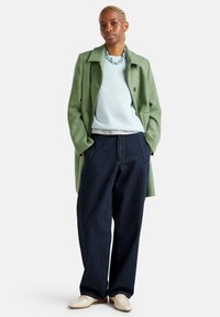 Persoon met gevlochten haar, in een lichtgroene jas, een lichtblauwe trui, donkere wijdvallende jeans, witte loafers en een stevige kettinghalsketting.