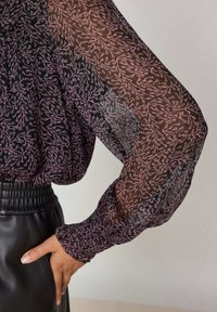 Blouse noire transparente avec motif floral rose, manches longues bouffantes et poignet élastique. Associée à une ceinture noire en faux cuir.