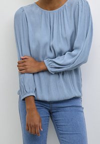 Blouse bleu clair, texturée, avec un col rond et des manches longues à volants. Le motif comprend de petits points en relief. Associée à un jean ajusté.
