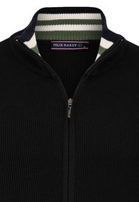 Sort strikket zip-up sweater med høj krave, der har en stribet indvendig foring i grøn og hvid samt en sølvfarvet lynlås. Mærket siger "Felix Hardy."