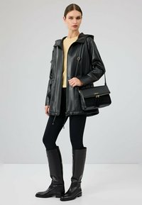 Giacca nera in pelle con cappuccio e chiusura con zip, indossata sopra una canotta giallo chiaro e leggings neri. Completata con stivali neri al ginocchio.