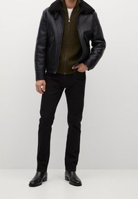 Giacca bomber in pelle nera con colletto in pelliccia, strato interno in maglia verde, pantaloni neri e stivaletti neri. Texture liscia, design aderente.