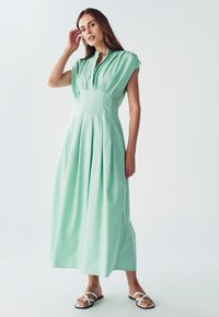 Mäta zelená maxi šaty s krátkymi, nariasenými rukávmi, zapínaním na gombíky na dekolte a plisovanou sukňou. Nosené s bielymi sandálmi na remienky.