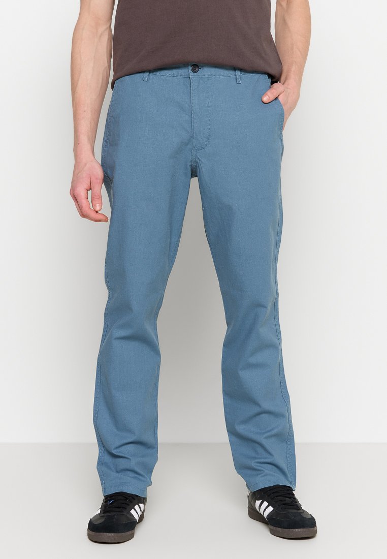 Chinos bleu clair en mélange de coton, avec une coupe classique, des poches avant et une fermeture éclair avec un bouton. Portés avec des baskets noires.