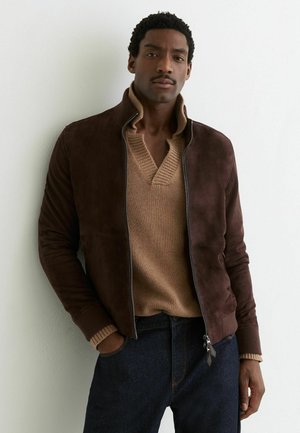 Jonge man met kort krullend haar en snor, gekleed in een bruine suède jas over een beige gebreide trui en donkere spijkerbroek, staande voor een effen witte muur.