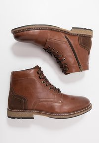 Botas de tobillo de cuero marrón con cordones en la parte delantera, cremallera en el costado y acentos de ante. Detalles de costura y suela de goma texturizada.