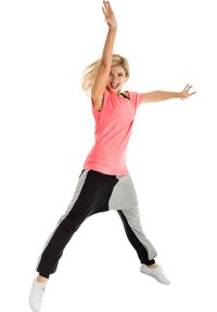 Roza majica s kratkimi rokavi in ohlapnim krojem, v kombinaciji z črnimi in sivo jogger hlačami z asimetričnim dizajnom. Model skače z dvignjenima rokama.