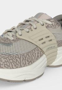 Κοντινή λήψη μπεζ και γκρι αθλητικού παπουτσιού με σχεδιασμένες επενδύσεις, πλέγμα στα πάνελ, χοντρή λευκή σόλα και λογότυπο "Jordan" στην πλευρική στήριξη.