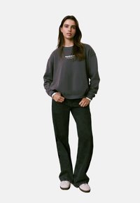 Sweatshirt gris avec un col rond et du texte brodé, associé à un jean large noir et des baskets blanches, sur un fond uni.
