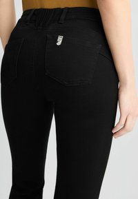 Zwarte jeans met een gladde textuur, voorzien van een hoge tailleband en achterzakken. Zilverkleurige accessoires accentueren de bovenste zak.