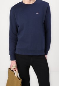 Person med en marinblå sweatshirt och svarta byxor, håller en tanfärgad canvasväska vid handtagen vid sin sida.