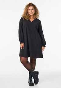 Zwarte jurk met een V-hals, lange mouwen met geplooide manchetten en een uitlopende vorm. Draag het met zwarte enkellaarzen en doorschijnende panty's.