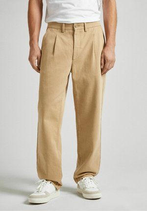 Homme portant un pantalon beige droit, des baskets blanches et une chemise blanche sur un fond clair uni.