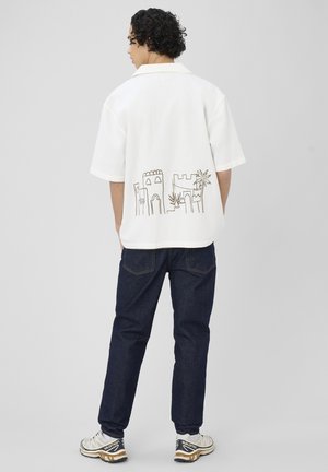 Homme aux cheveux bouclés portant une chemise blanche à manches courtes avec un dessin en lignes noires représentant un château et des palmiers, un jean foncé et des baskets blanches.