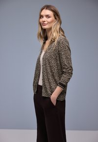 Blouson aviateur à imprimé léopard en beige et noir avec poignets côtelés, associé à un haut clair et un pantalon noir. Tissu lisse, coupe décontractée.
