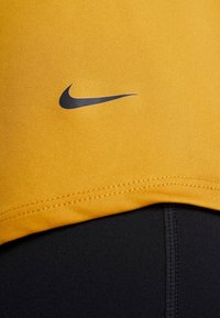 Canotta sportiva gialla realizzata in tessuto liscio con il logo swoosh nero di Nike e leggings neri visibili nella parte inferiore, caratterizzati da texture contrastanti.