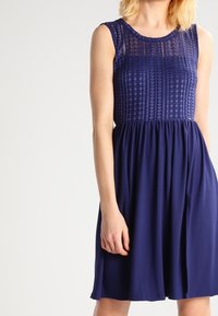 Navy-blaues ärmelloses Kleid mit einem strukturierten Oberteil mit geometrischen Mustern und einem fließenden, gerafften Rock. Weicher Stoff mit leichtem Schimmer.