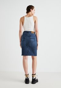 Denim kokerrok met achterzakken, een lichte taper en een Lee-label. Gedragen met een witte tanktop en zwarte enkellaarsjes met beige accenten.