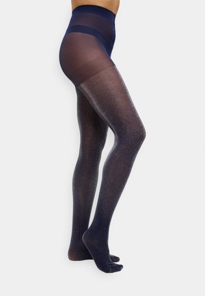 Noge u visokim, prozirnim tamnoplave čarape s glitter efektom od struka do prstiju, stojeće protiv jednostavne bijele pozadine.