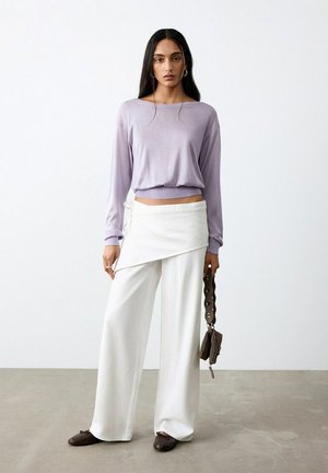 Kobieta ubrana w fioletowy top z długim rękawem, białe szerokie spodnie z detalem wiązania, brązowe baleriny i trzymająca brązową torebkę z pętlą na pasku.